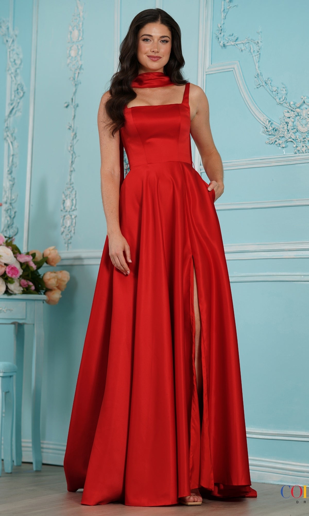 A woman wears a red Colors Dress 4157 floor-length satin A-line gown featuring a square neckline, a bow sash, a side slit, and pockets.